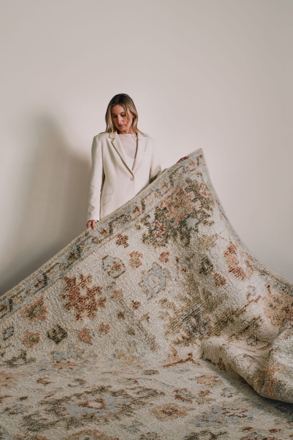 Woman in a white suit holding the Hemen rug from the Oushak Collection, featuring a traditional pattern in cream, soft blue, olive and terracotta.